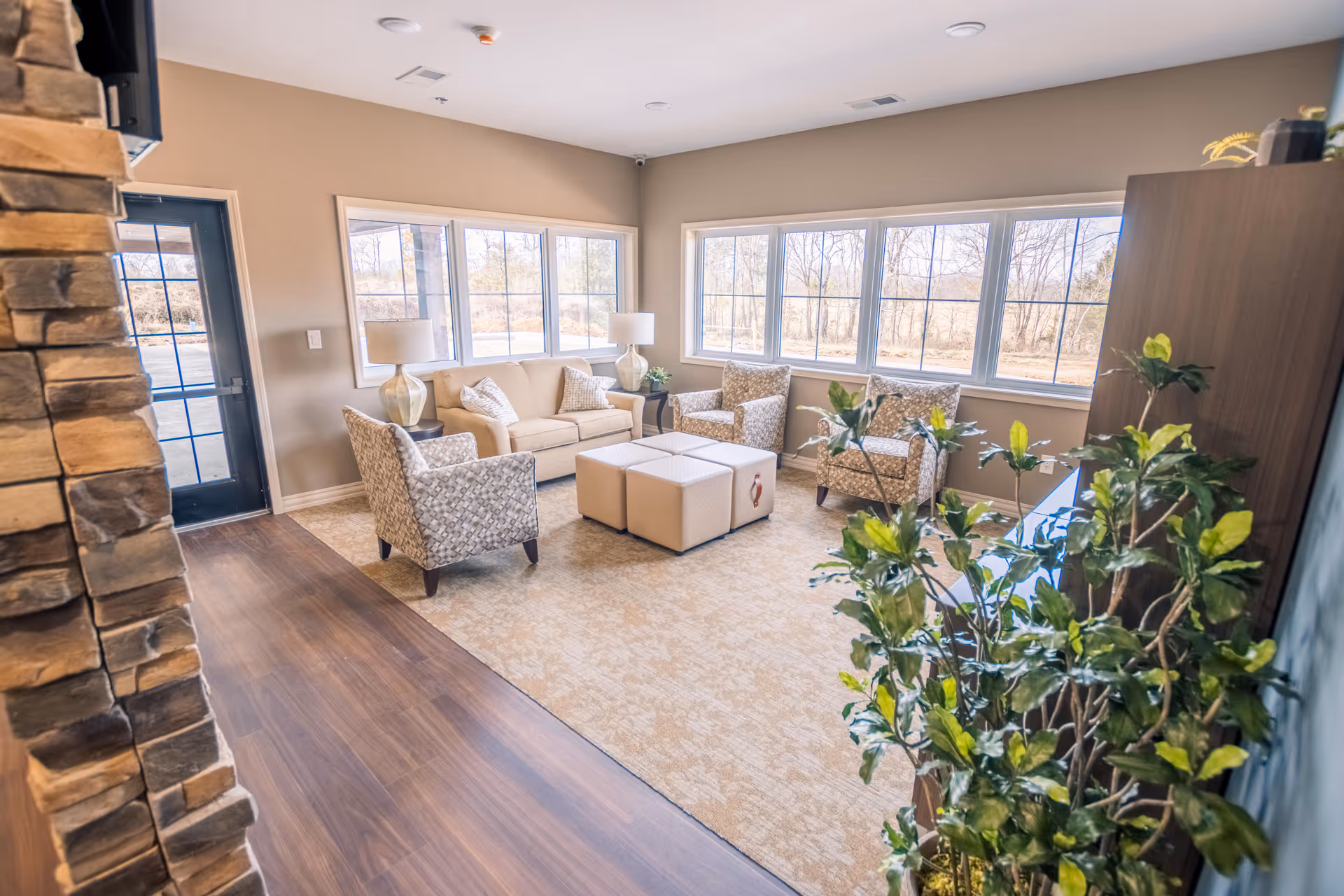 Bright living room lounge with beige sofas, patterned chairs, ottomans, lamps, and large windows.