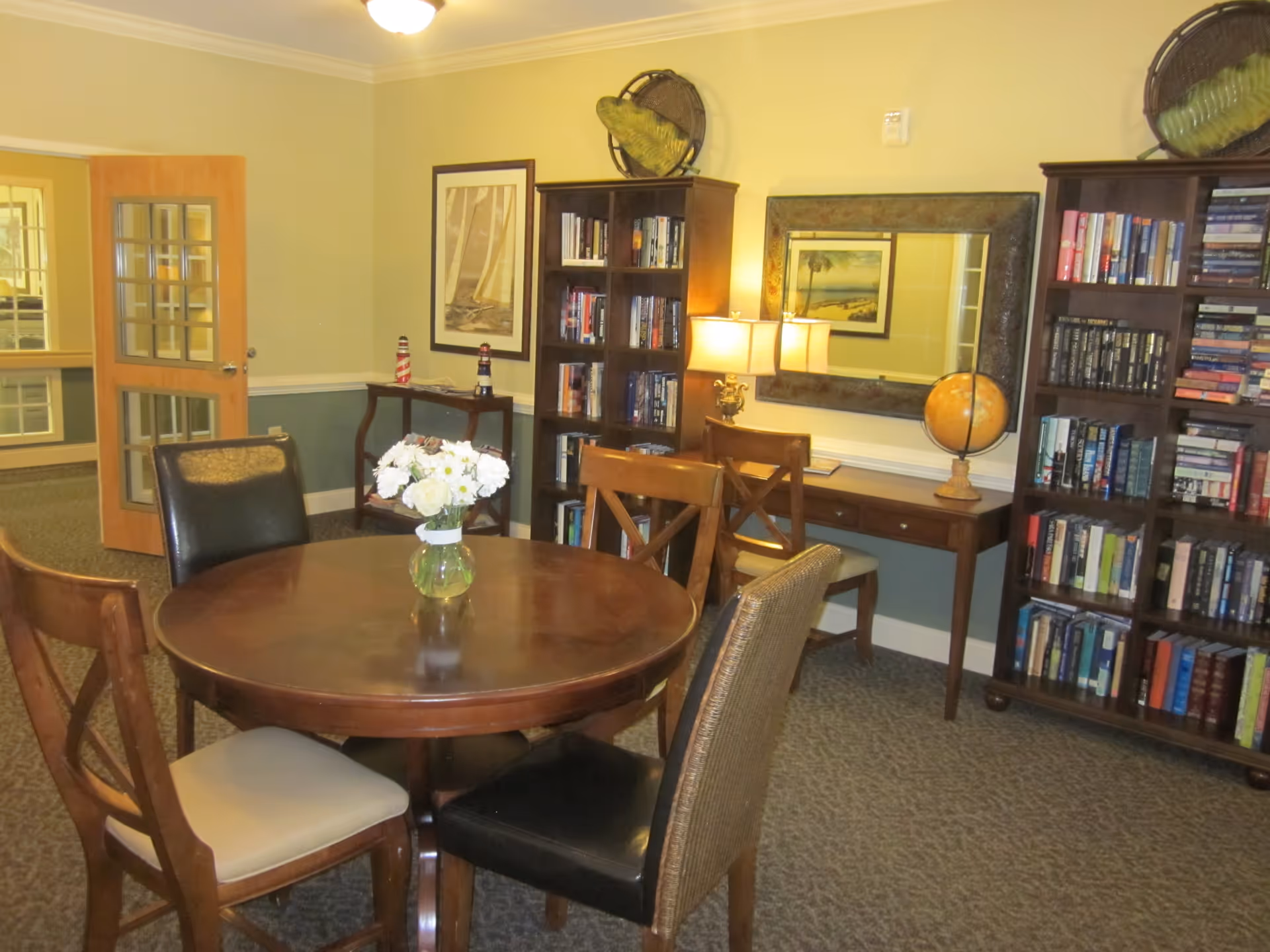 A cozy interior room with a round wooden table surrounded by four chairs, a vase with white flowers on the table, two tall bookshelves filled with books, a wooden desk with a globe and a lamp, a large mirror on the wall, and framed artwork. The room has beige walls and carpeted floor.