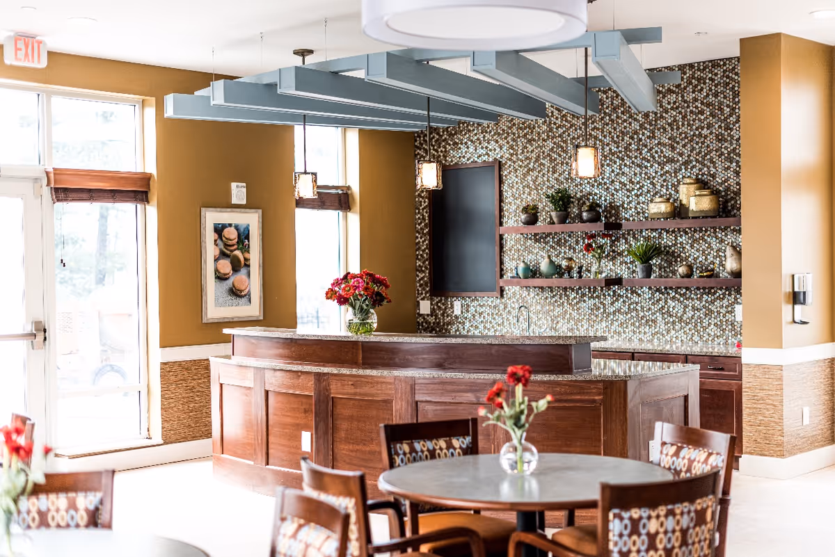Bright communal dining area with round tables and patterned chairs facing a wooden service counter with a mosaic tile backsplash and decorative shelves.