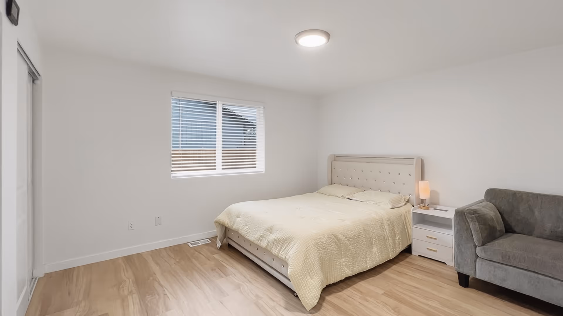 Bright bedroom with large bed, nightstand, lamp, and gray sofa.