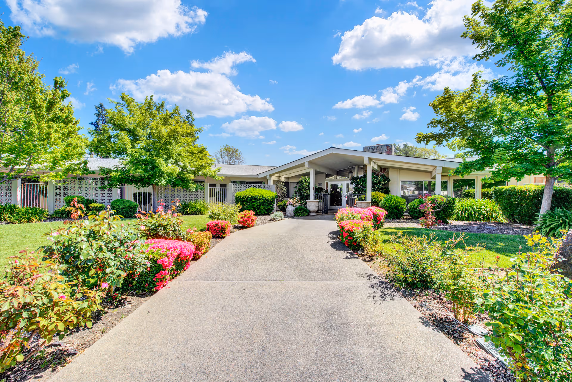 A single-story building with a covered entrance surrounded by well-maintained gardens and colorful flowering bushes under a bright blue sky with scattered clouds.