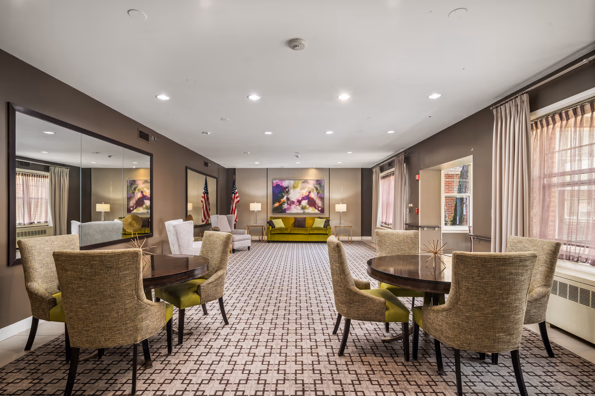 Well-lit senior living lounge with round tables, upholstered chairs, and artwork.
