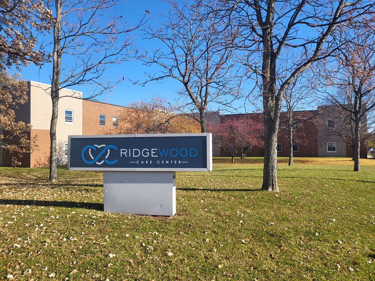 Outdoor view of Ridgewood Care Center sign on a grassy area with trees and a brick building in the background under a clear blue sky.