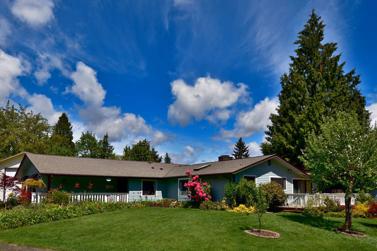 Single-story ranch-style house with a large green lawn, flowering shrubs, and tall trees under a blue sky.