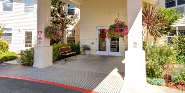 Entrance area of a senior living facility with a covered walkway supported by columns, hanging flower baskets, a bench, and glass double doors leading inside. The exterior walls are light-colored with windows and surrounding greenery including bushes and small trees.