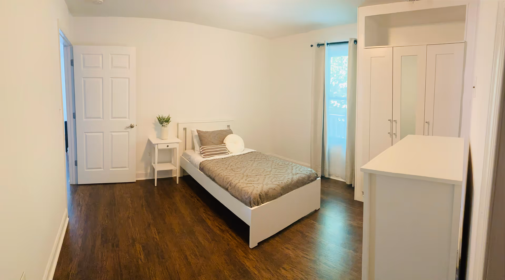 A simple, clean bedroom with a single bed covered in a gray patterned blanket and pillows. There is a small white nightstand with a potted plant next to the bed. The room has wooden flooring, a white wardrobe with mirrored doors, a white dresser, and a window with light curtains allowing natural light to enter.