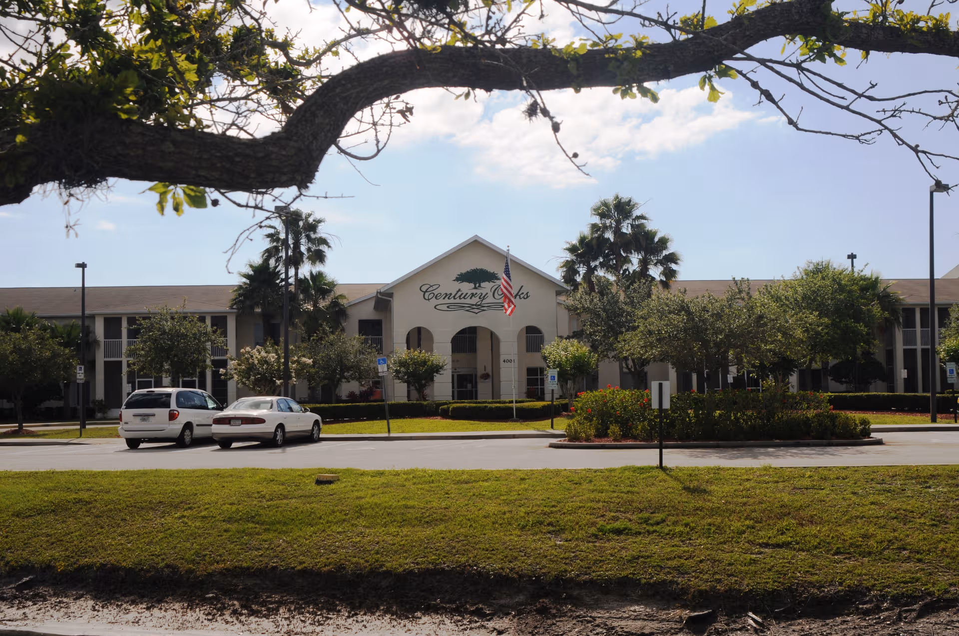 Front view of Century Oaks senior living facility with parking, trees, and a flag.