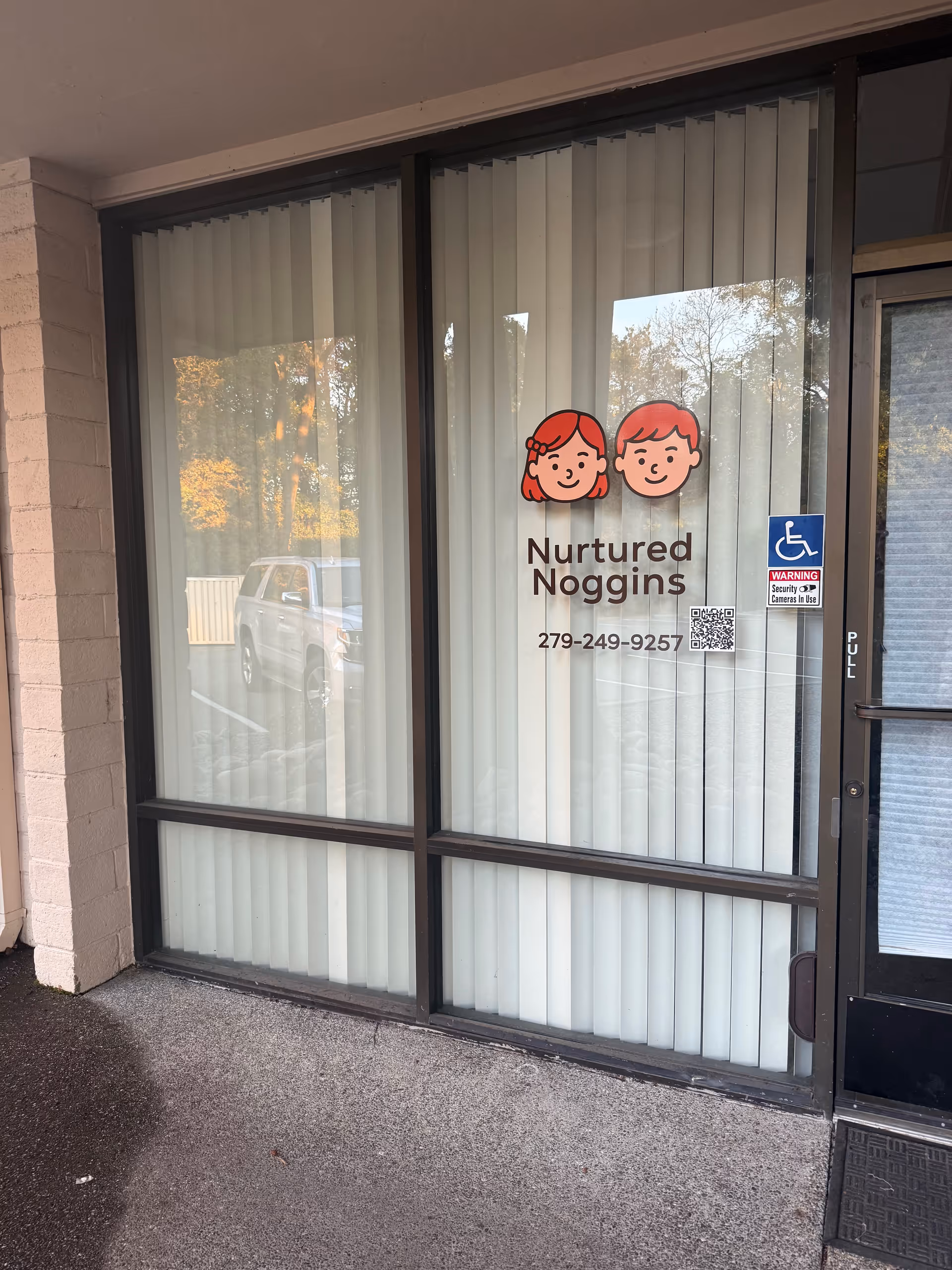 Glass storefront entrance with Nurtured Noggins sign, cartoon logo, and accessibility sticker.