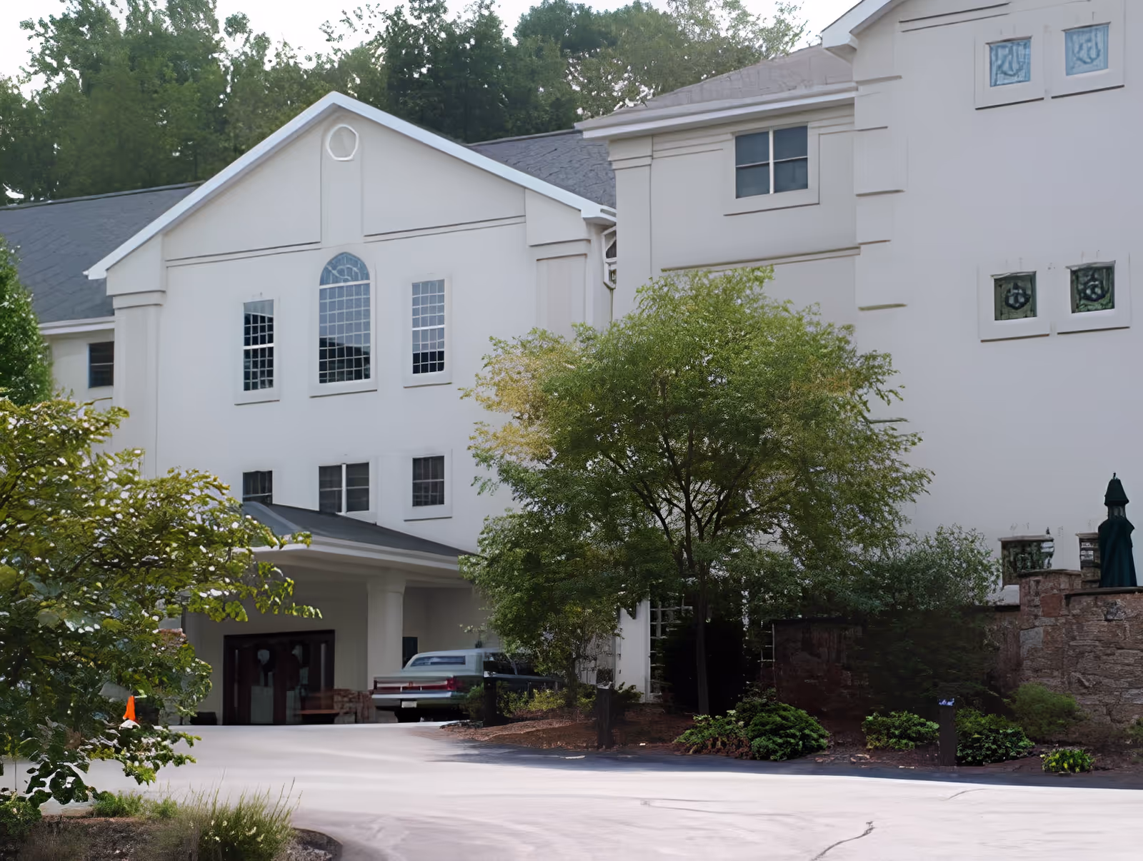 Exterior view of a large white building with multiple windows, surrounded by trees and greenery. A driveway leads to a covered entrance where a car is parked.