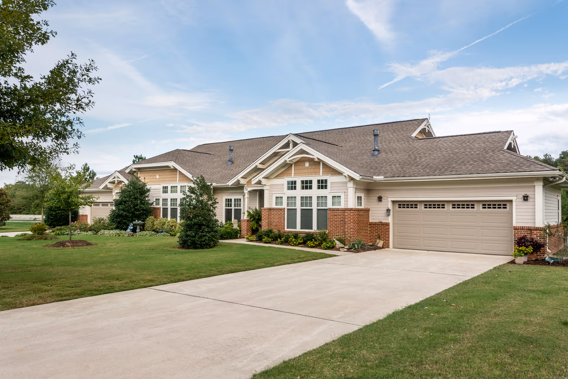Exterior view of a single-story residential building with a large driveway, a two-car garage, and well-maintained landscaping including trees and shrubs under a partly cloudy sky.
