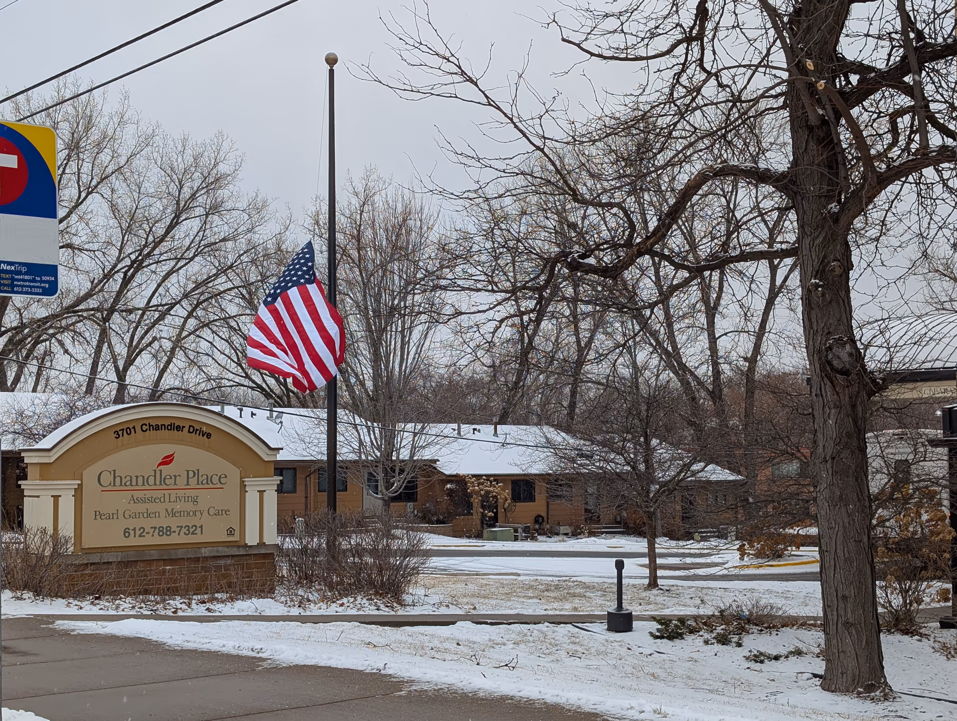 Exterior view of an assisted living facility sign and building with an American flag at half-mast in a snowy yard.
