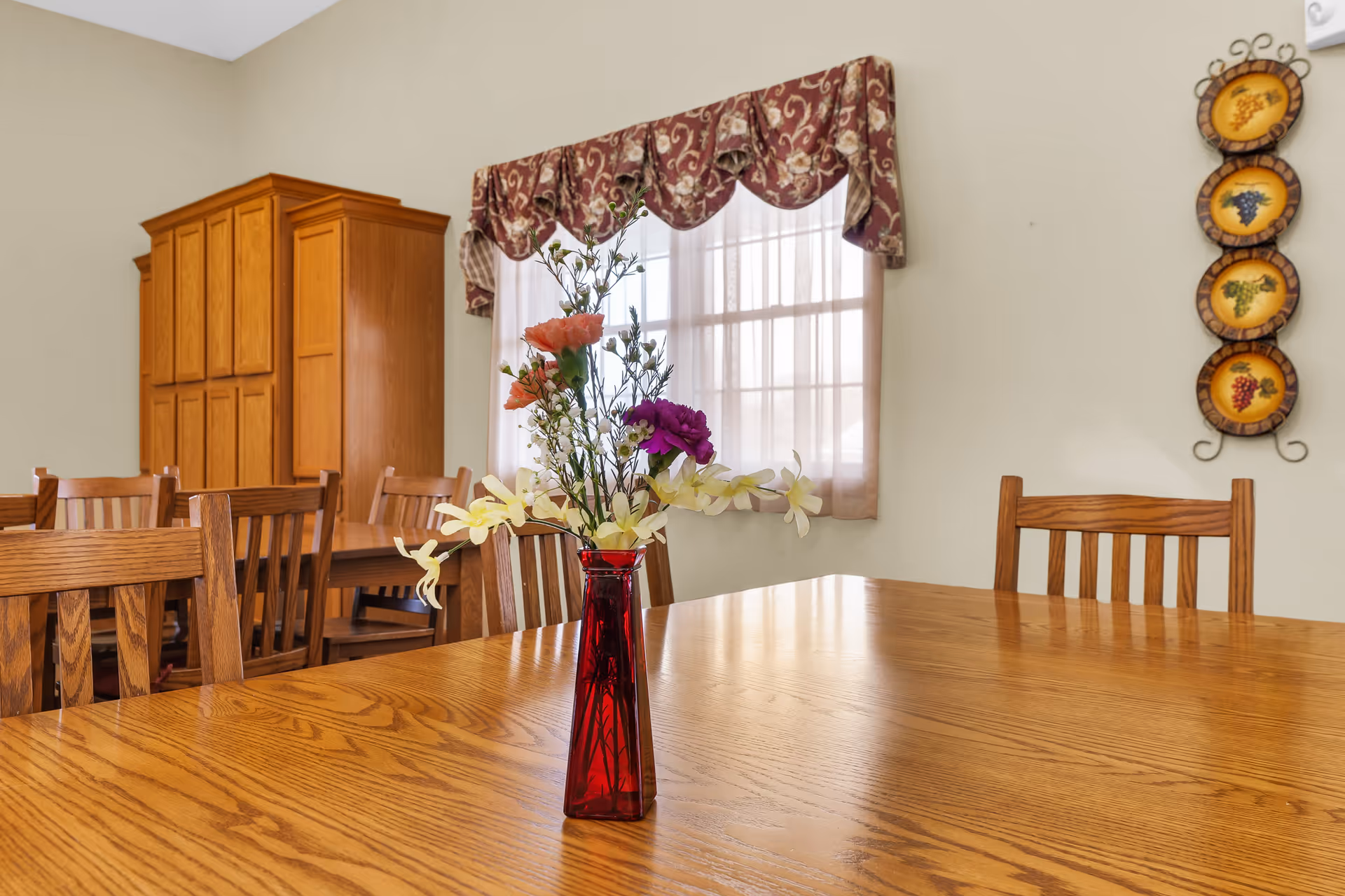 Wooden dining table with a red vase of flowers in a bright, cozy dining room.