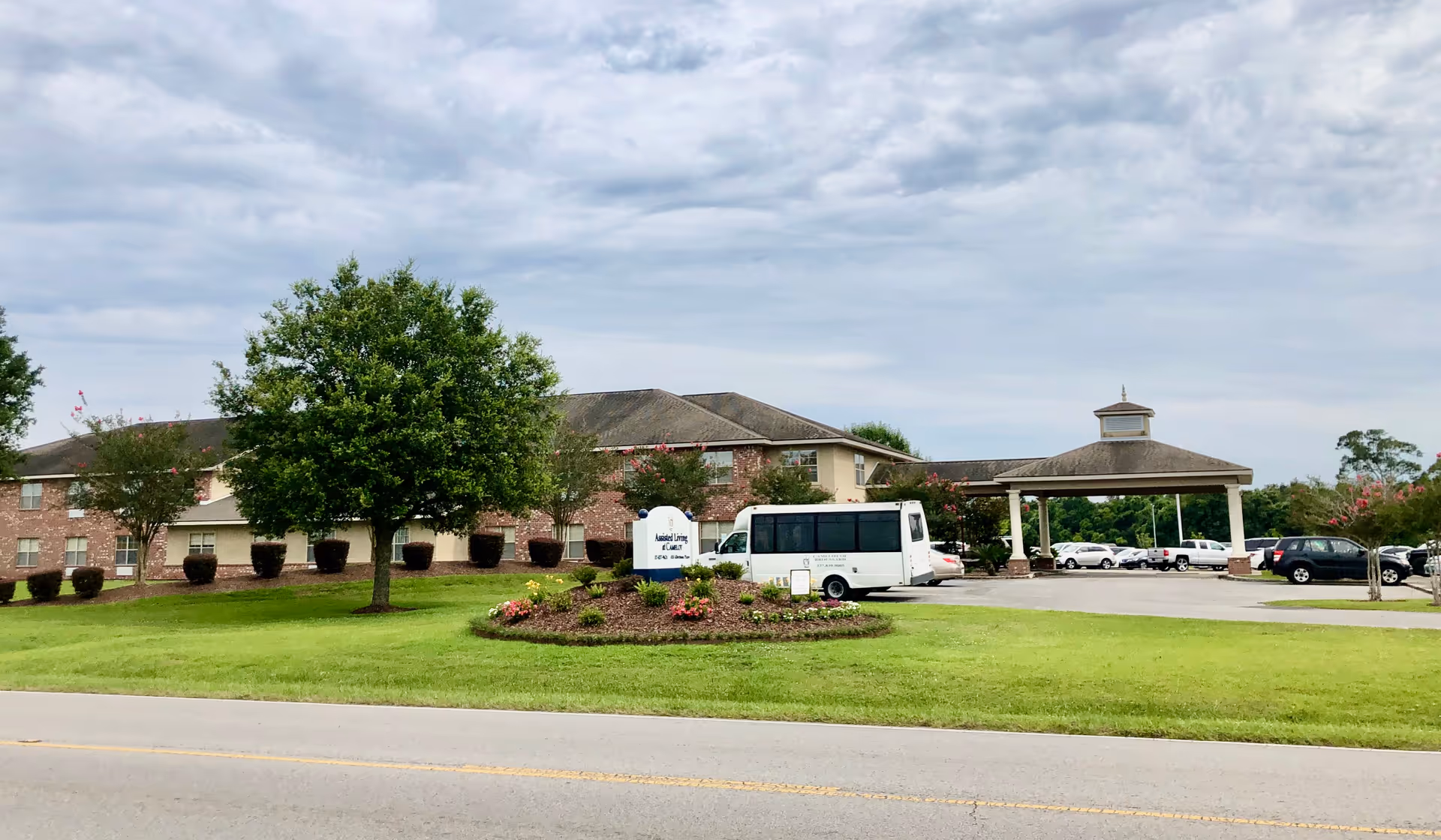 Exterior view of Camelot of Broussard showing the entrance canopy, lawn, and parked bus.