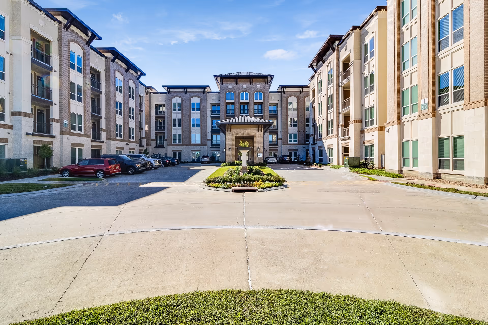 Exterior view of a multi-story residential building with a central entrance and a small landscaped area with a fountain in front. Several cars are parked along the driveway on either side of the entrance under a clear blue sky.