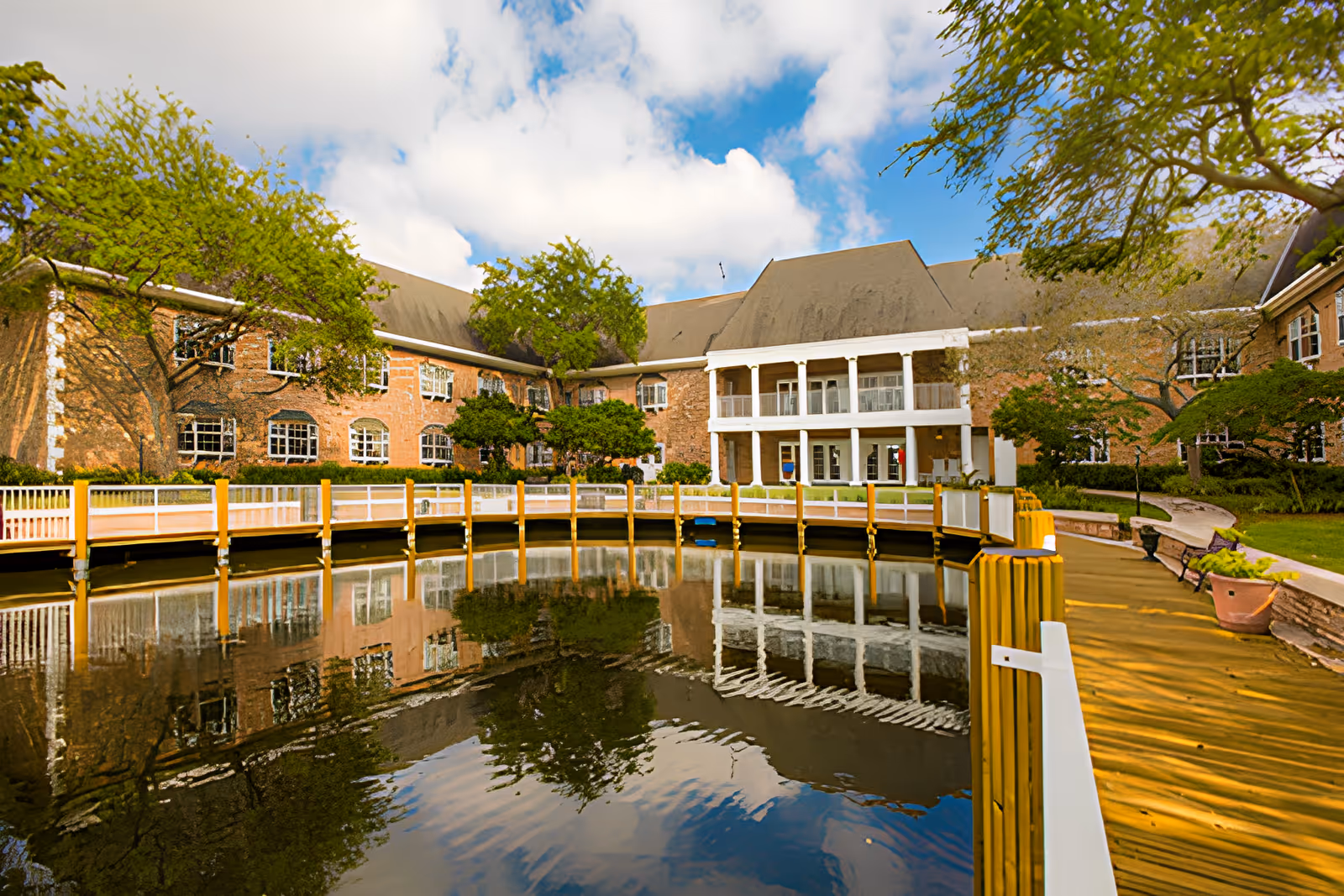 Brick assisted-living building with a white-columned porch reflected in a pond and a wooden boardwalk in the foreground.