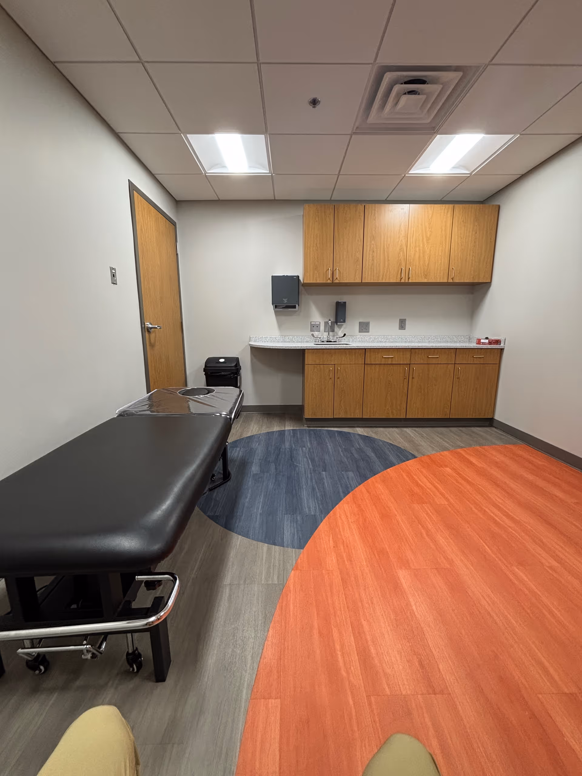Clinical exam room with a black examination table, cabinets, sink, and colorful circular floor patterns.