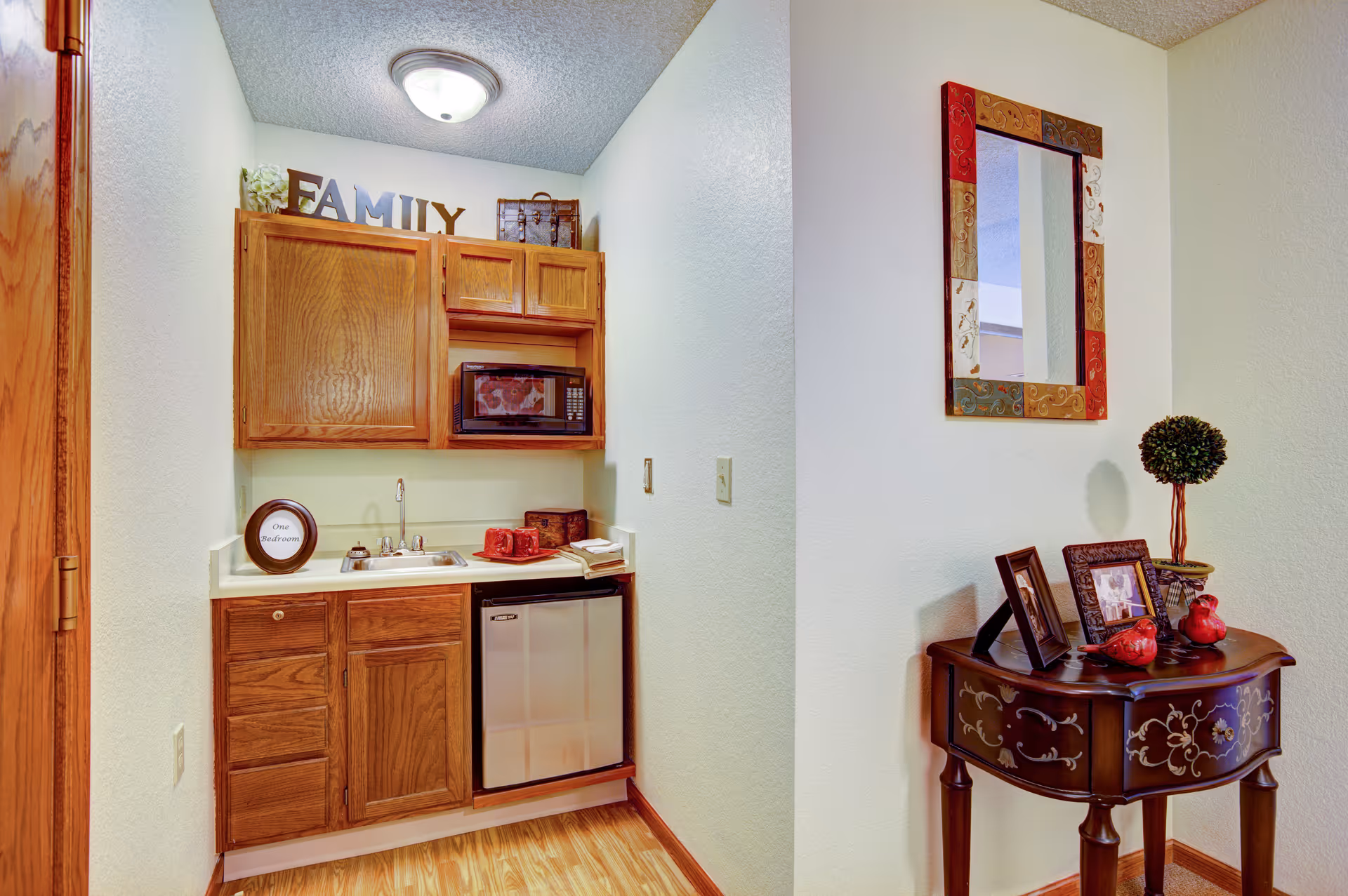 Wooden kitchenette with cabinets, sink, microwave, mini-fridge, and decorative hallway table.