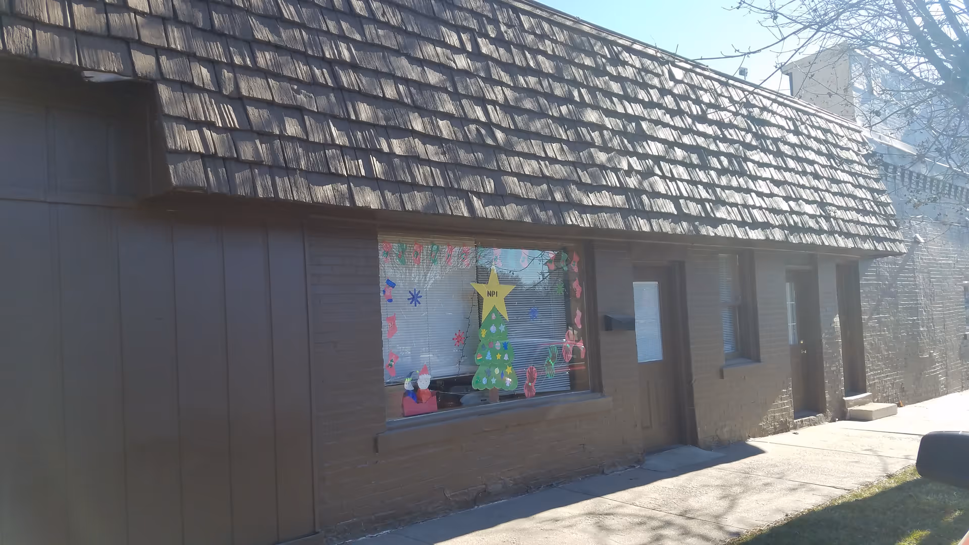 Brick exterior building with a decorated window and Christmas tree display under a shingled roof.