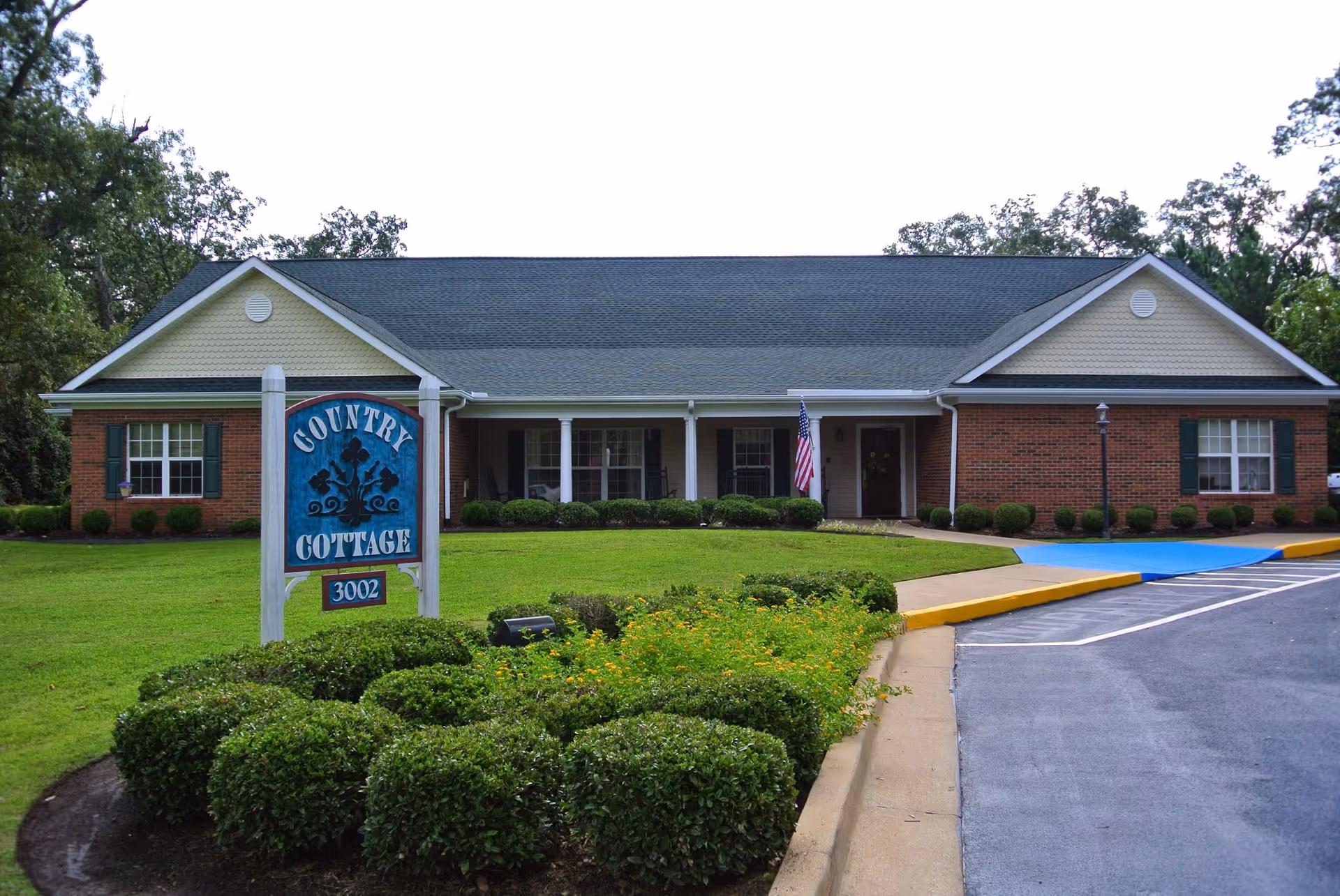 Front exterior view of a single-story brick building with a gray roof, green shutters, and a porch with white columns. There is a blue sign in front that reads 'Country Cottage 3002' surrounded by neatly trimmed bushes and a well-maintained lawn. An American flag is displayed near the entrance, and there is a parking area with a blue handicap ramp.