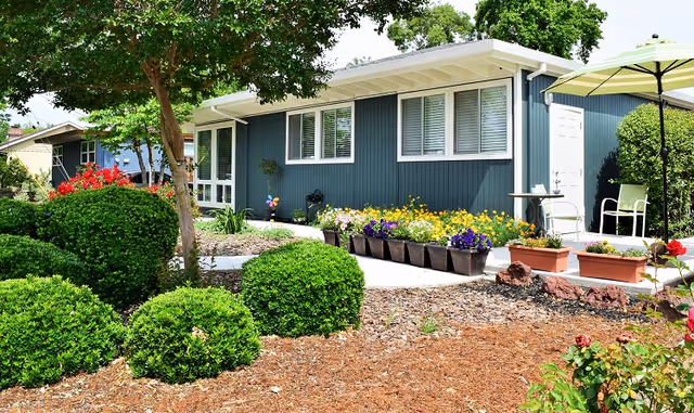 Blue single-story senior living exterior with colorful flower planters and a shaded patio.