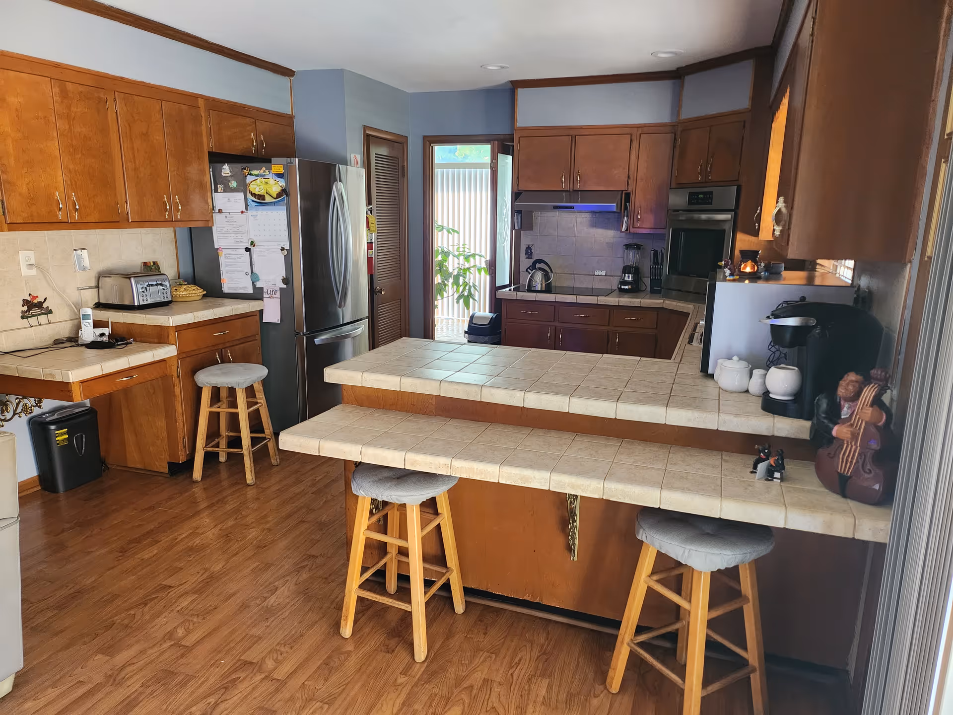 Spacious kitchen with wooden cabinets, tile counters, stainless appliances, and three bar stools.