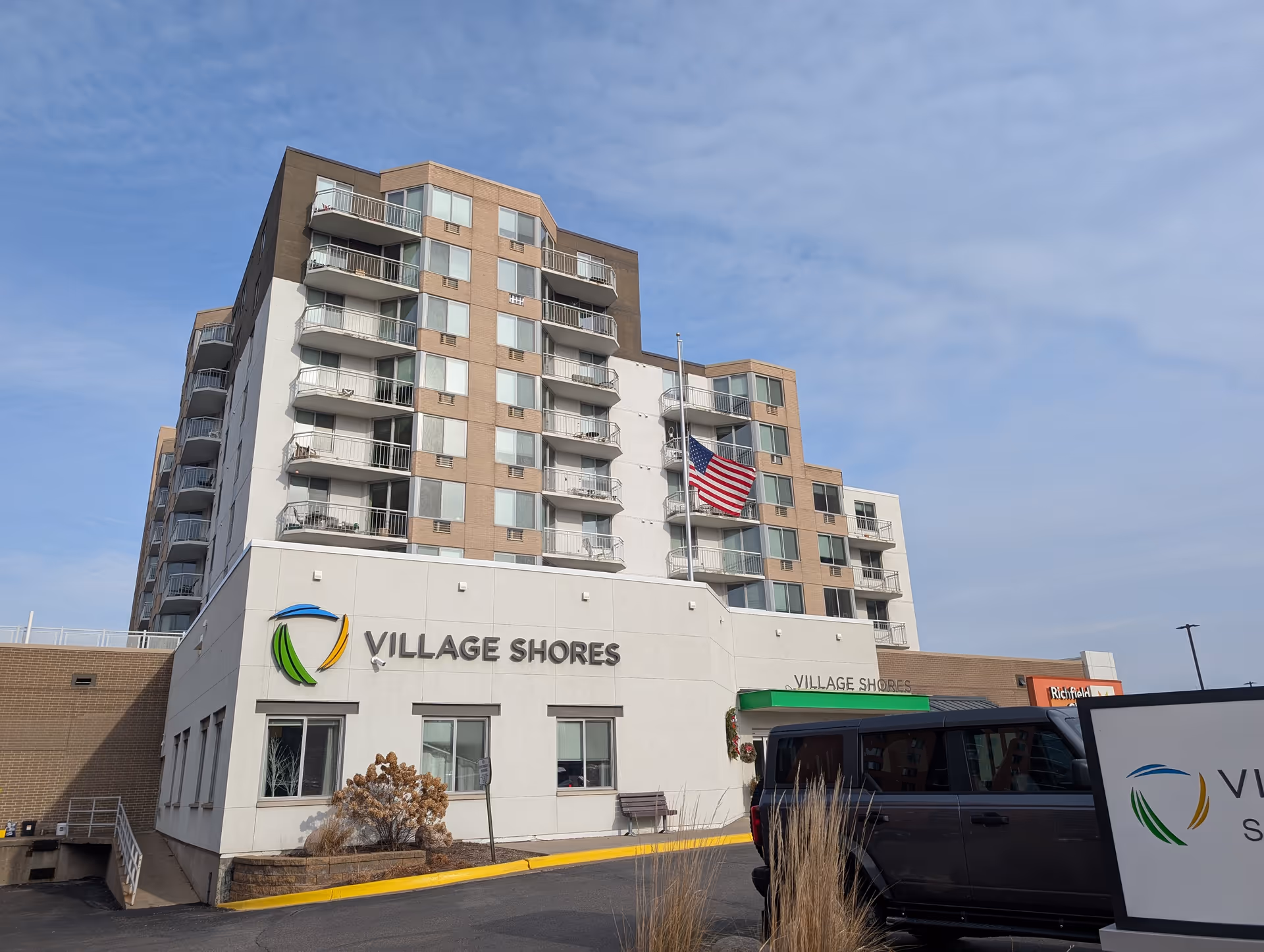 Exterior view of the Village Shores senior living facility building with multiple floors and balconies, an American flag on a flagpole, a parked black vehicle, and a clear sky.