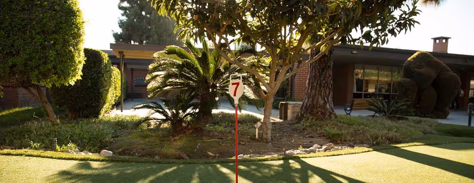 Outdoor garden area at Rowntree Gardens featuring trimmed bushes, palm plants, a putting green with a flag marked number 7, and a brick building in the background.