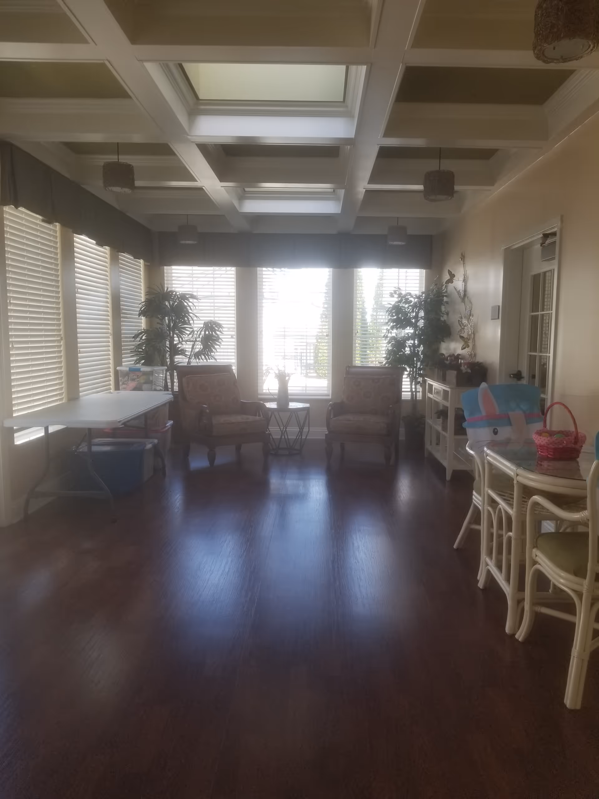 Bright senior living lounge with two armchairs, tables, plants, and large windows.