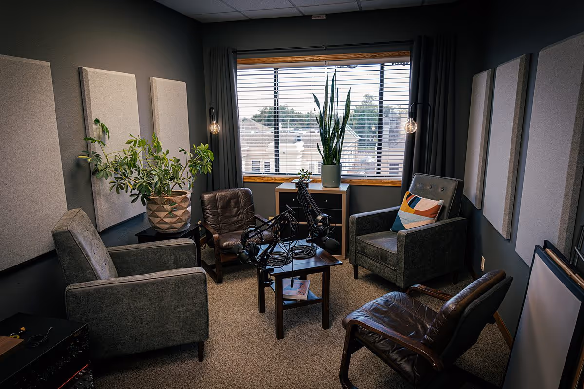 Cozy interior recording lounge with chairs, plants, microphones, and a large window.