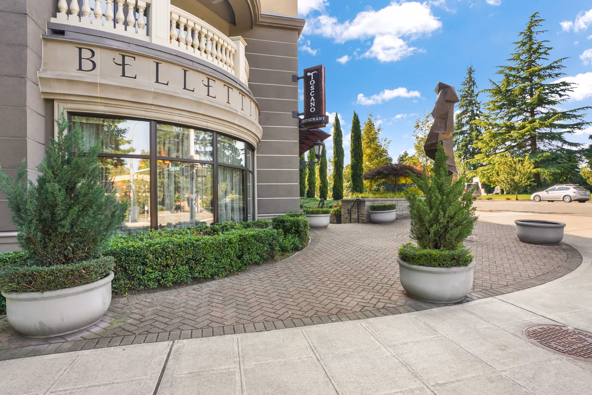 Exterior view of The Bellettini building showing a curved wall with large windows and the name 'BELLETTINI' displayed above. There are large potted plants along a brick-paved walkway leading to an outdoor area with trees, a sculpture, and a sign for Toscano Restaurant. A car is parked in the background under a blue sky with some clouds.