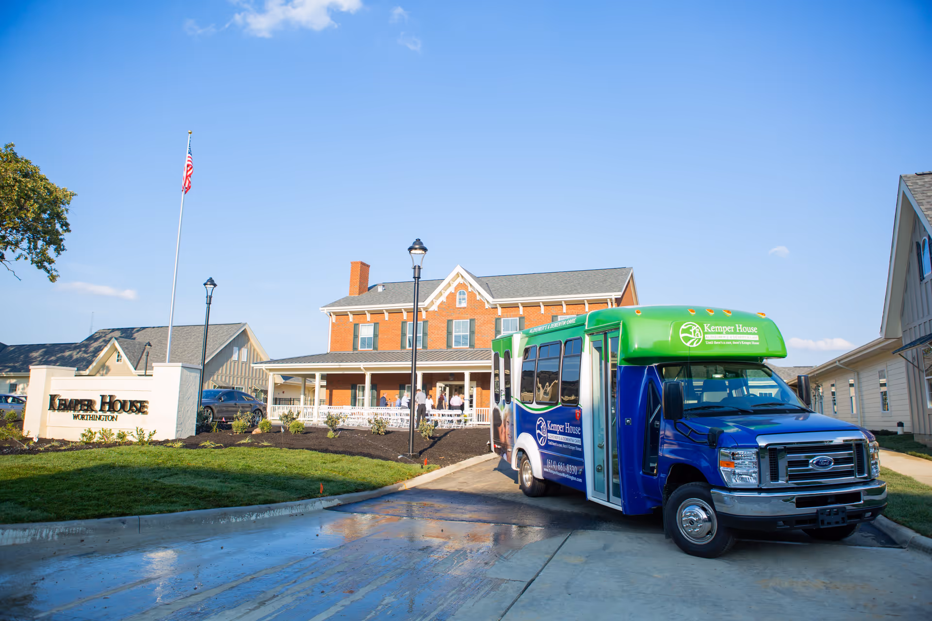 Exterior view of Kemper House Worthington, a senior living facility, showing a brick building with a porch and several people standing outside. A blue and green shuttle bus with Kemper House branding is parked in front. There is a sign with the facility name and an American flag on a flagpole nearby under a clear blue sky.