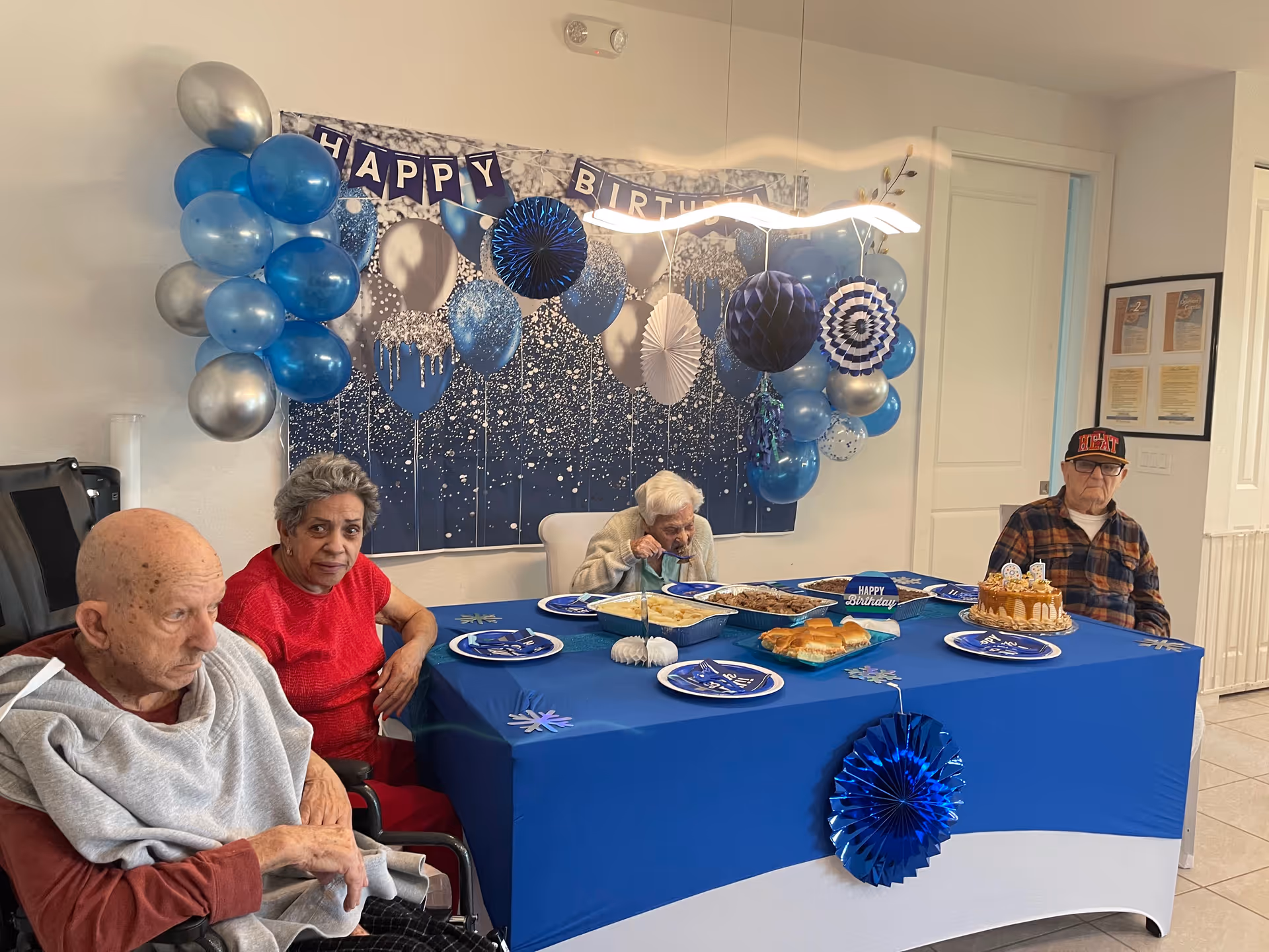 Four elderly individuals sitting around a table decorated with a blue tablecloth and birthday decorations including blue and silver balloons, a 'Happy Birthday' banner, and a birthday cake. The table has plates, food trays, and a small birthday sign. The setting appears to be a celebration in a senior living facility.