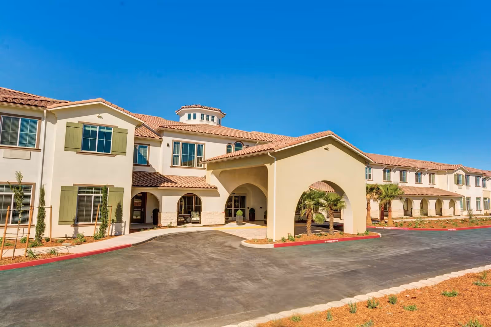Front exterior of a Mediterranean-style senior living facility with a covered arched entrance, landscaped grounds, and a driveway.