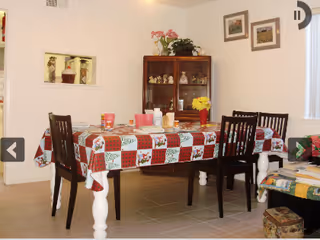 Dining room with a table covered in a red-and-white patchwork tablecloth, four chairs, and a china cabinet against a white wall.