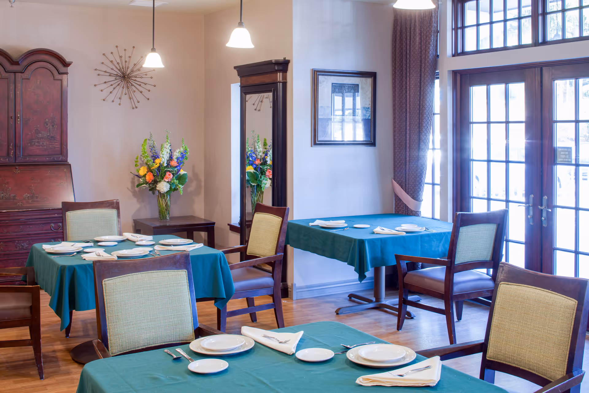 A dining room with several tables covered in teal tablecloths, each set with white plates, napkins, and silverware. The room features wooden chairs with light green cushions, a wooden cabinet, a wall-mounted decorative piece, a tall mirror, a vase with colorful flowers, and large windows with glass doors letting in natural light.