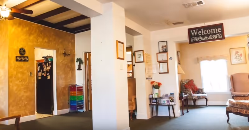 Bright communal living area with chairs, a "Welcome" sign, and open doorways to adjacent rooms.