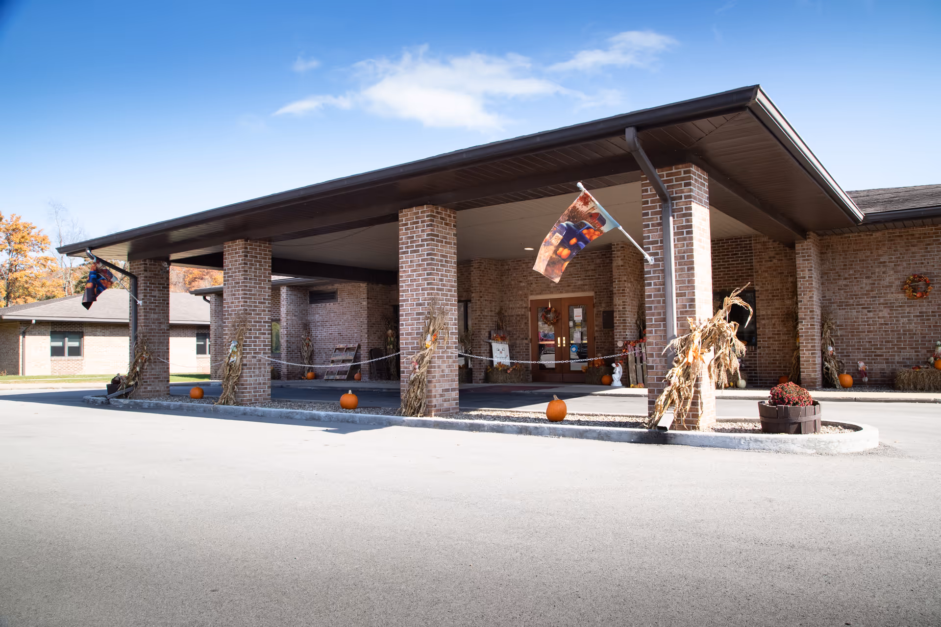 Brick building entrance with a covered drop-off, fall pumpkins, and corn stalks.
