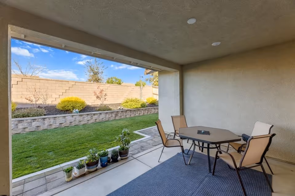 Covered outdoor patio with a table, chairs, and potted plants along a green lawn.