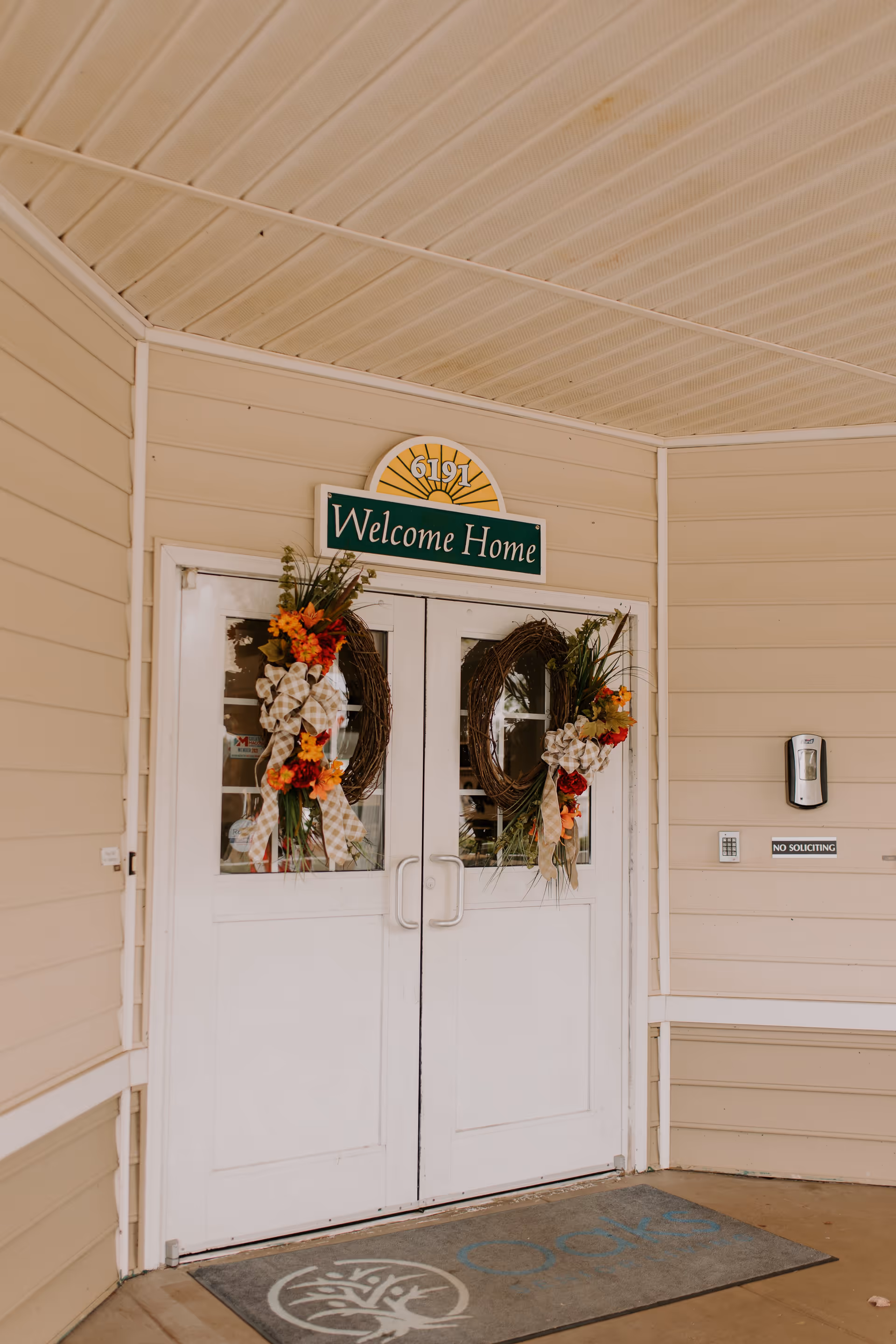 Double white doors decorated with autumn-themed wreaths featuring flowers and ribbons. Above the doors is a sign with the number 6191 and a green sign that reads 'Welcome Home.' To the right of the doors is a hand sanitizer dispenser and a 'No Soliciting' sign. A doormat with a tree logo is placed in front of the entrance.
