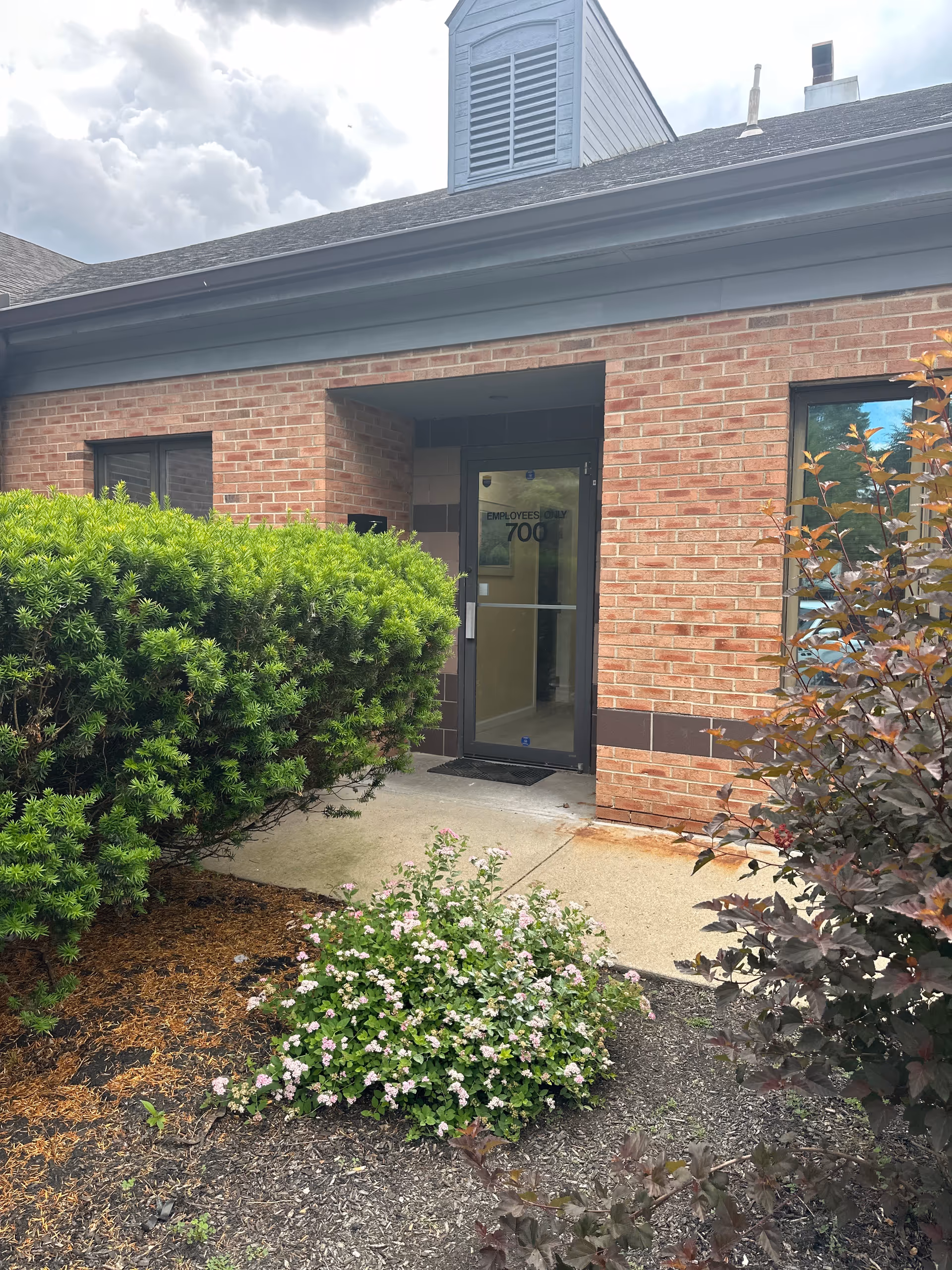 Brick facility entrance with glass door and surrounding shrubs and landscaping.