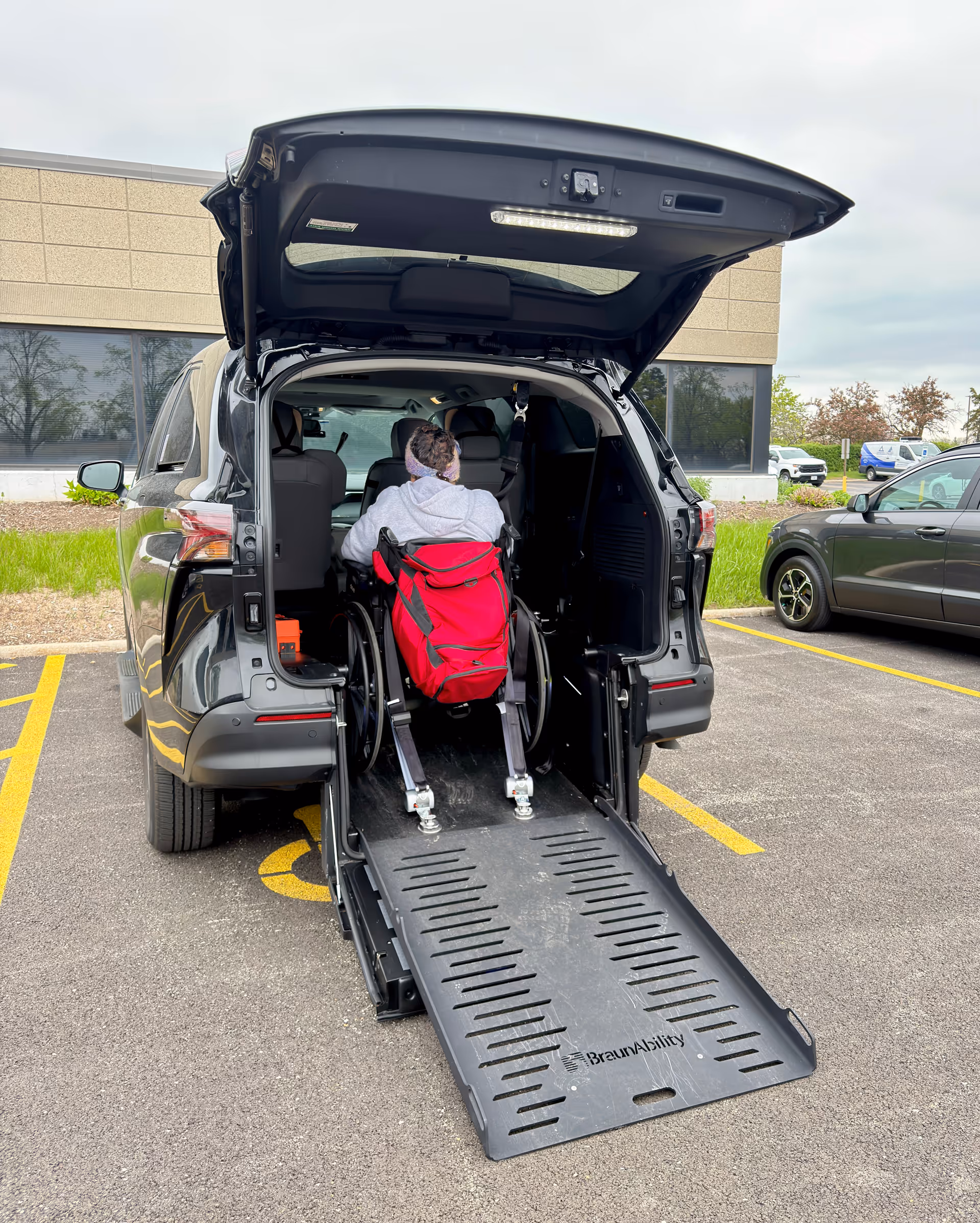 Wheelchair user in a red chair boards a vehicle via a ramp in a parking lot.