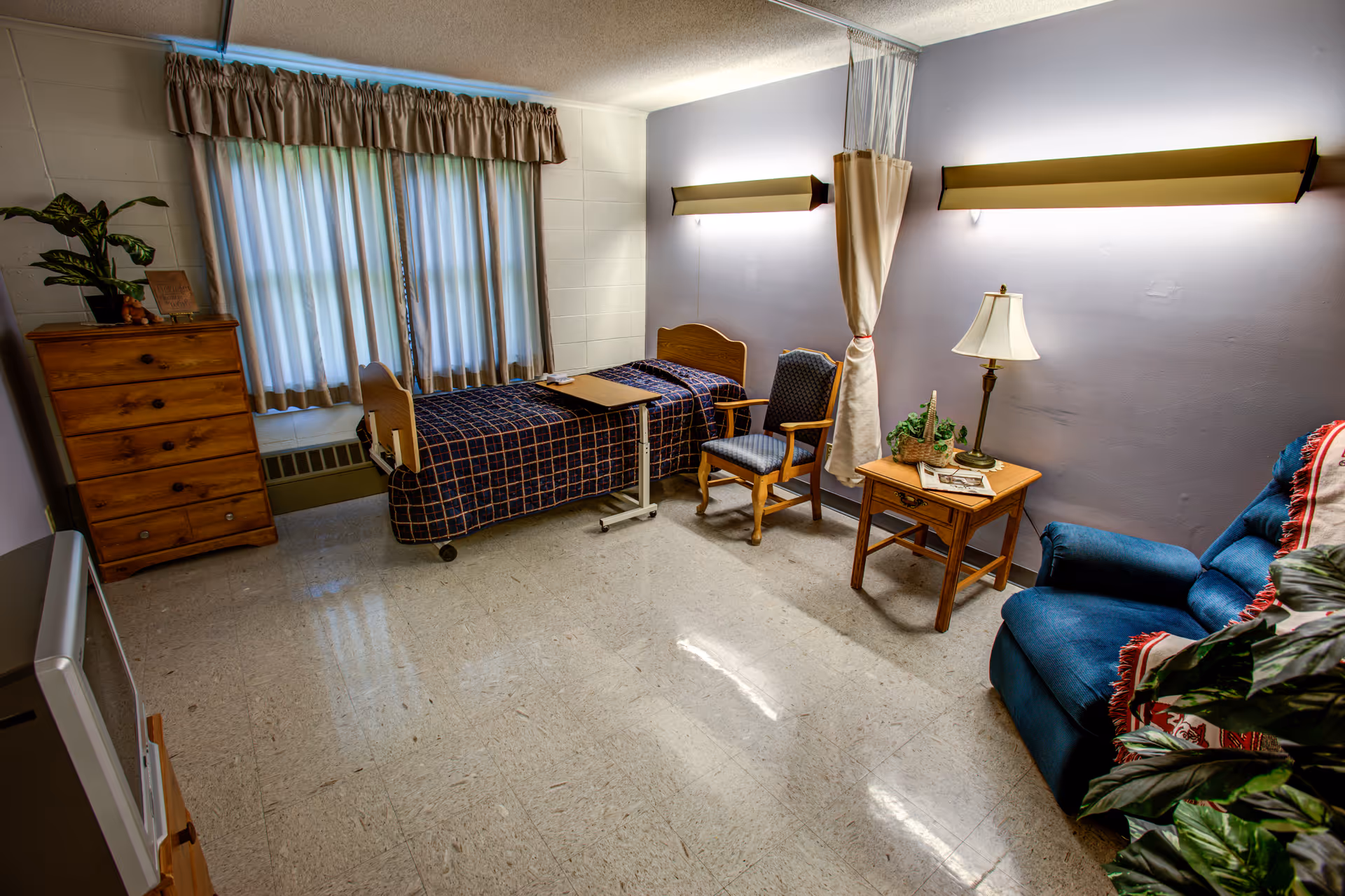 Small senior living bedroom with single bed, dresser, and blue seating area.