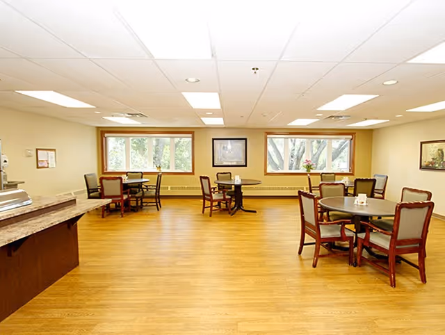 Bright dining room with round tables, chairs, and large windows in a senior facility.