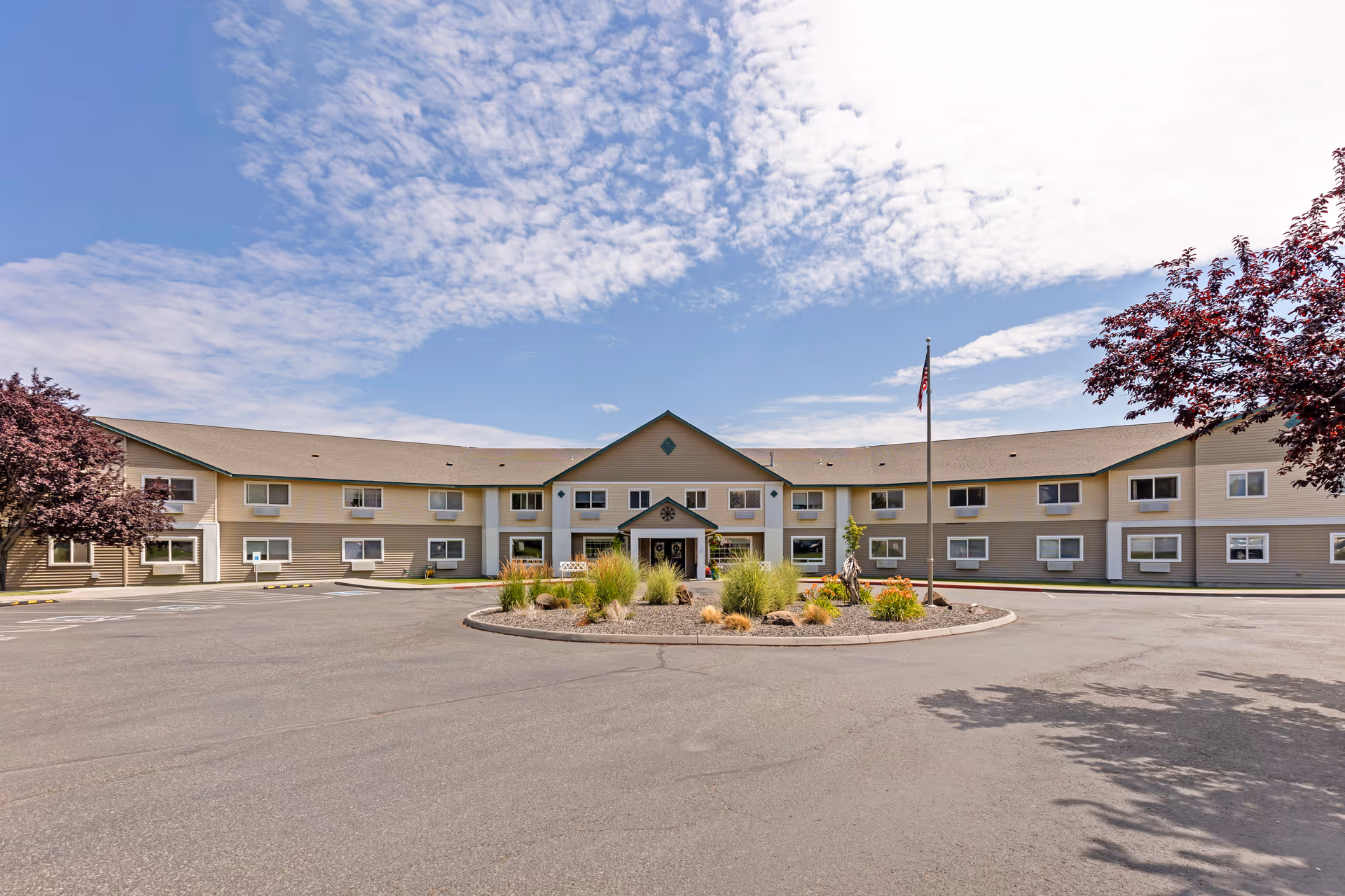 Front exterior view of Brookdale Meadow Springs facility showing a two-story building with beige siding, multiple windows, a central entrance with a small covered porch, a circular driveway with landscaped plants in the center, an American flag on a flagpole, and trees on either side under a partly cloudy sky.