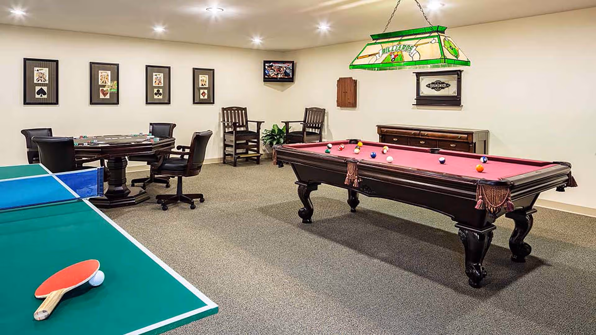 A recreational room with a green and blue ping pong table with a paddle and ball, a poker table with chairs, a pool table with balls arranged on it, two wooden chairs, a small TV mounted on the wall, and framed playing card artwork on the wall.