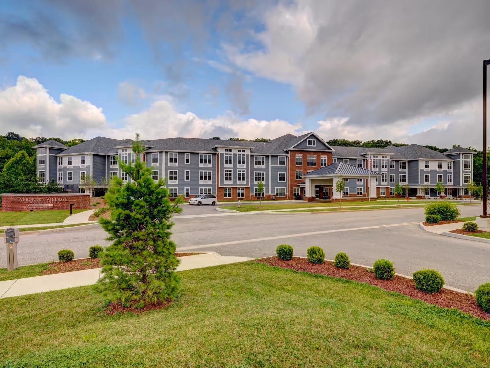 Exterior view of Evergreen Village at Bloomington, a large multi-story senior living facility with a combination of gray and brick facade, surrounded by landscaped greenery and a driveway in front under a partly cloudy sky.