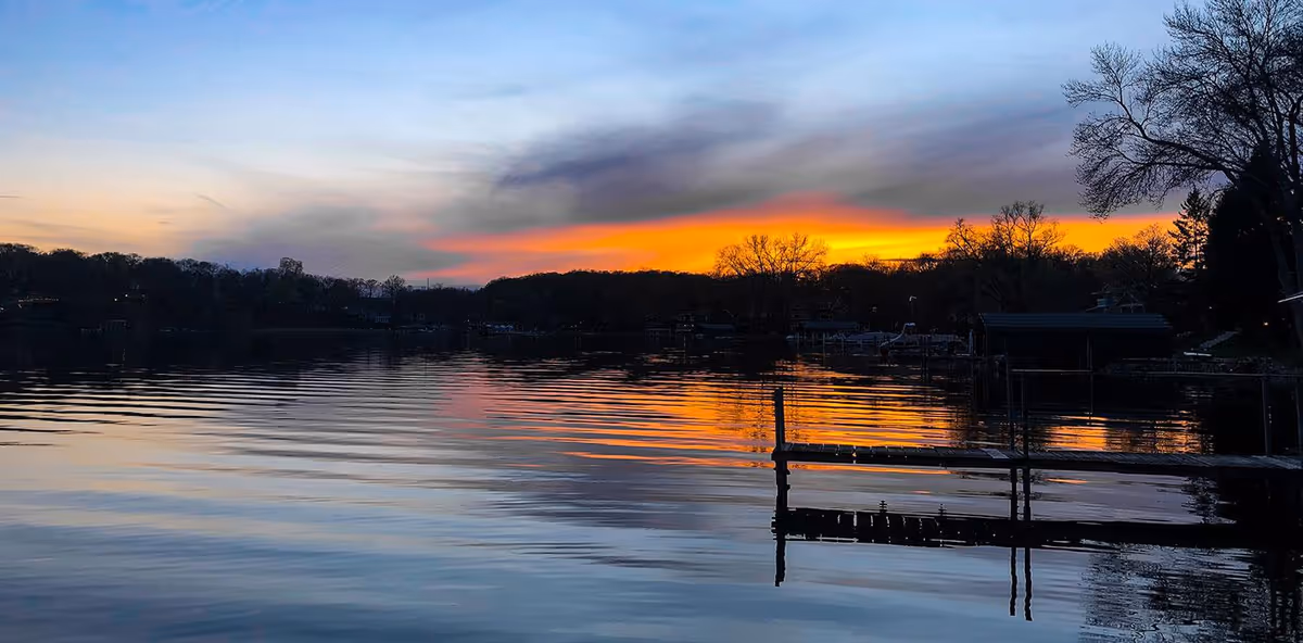 Sunset over a calm lake with a wooden dock and silhouetted trees along the shore.