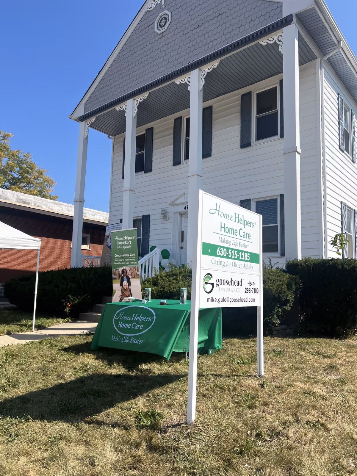 Two-story white building with decorative trim, signs, and a green table outside.