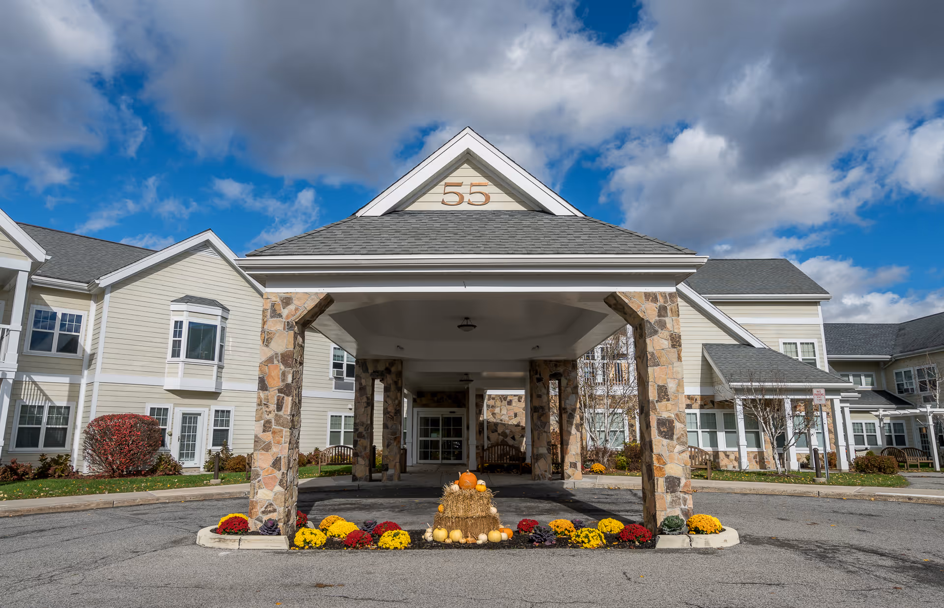 Front entrance and porte-cochère of a senior living building with stone columns, seasonal flowers and pumpkins under a partly cloudy sky.
