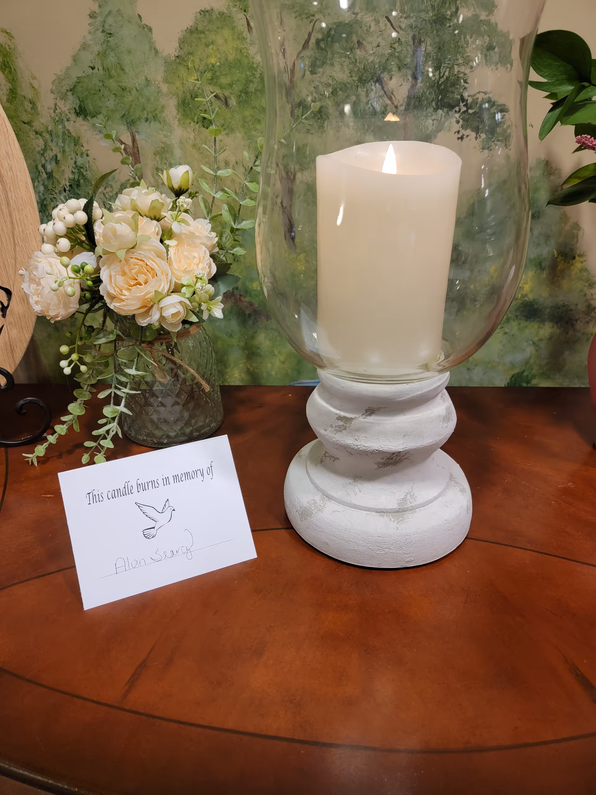 Lit candle inside a glass hurricane with flowers on a wooden table indoors.